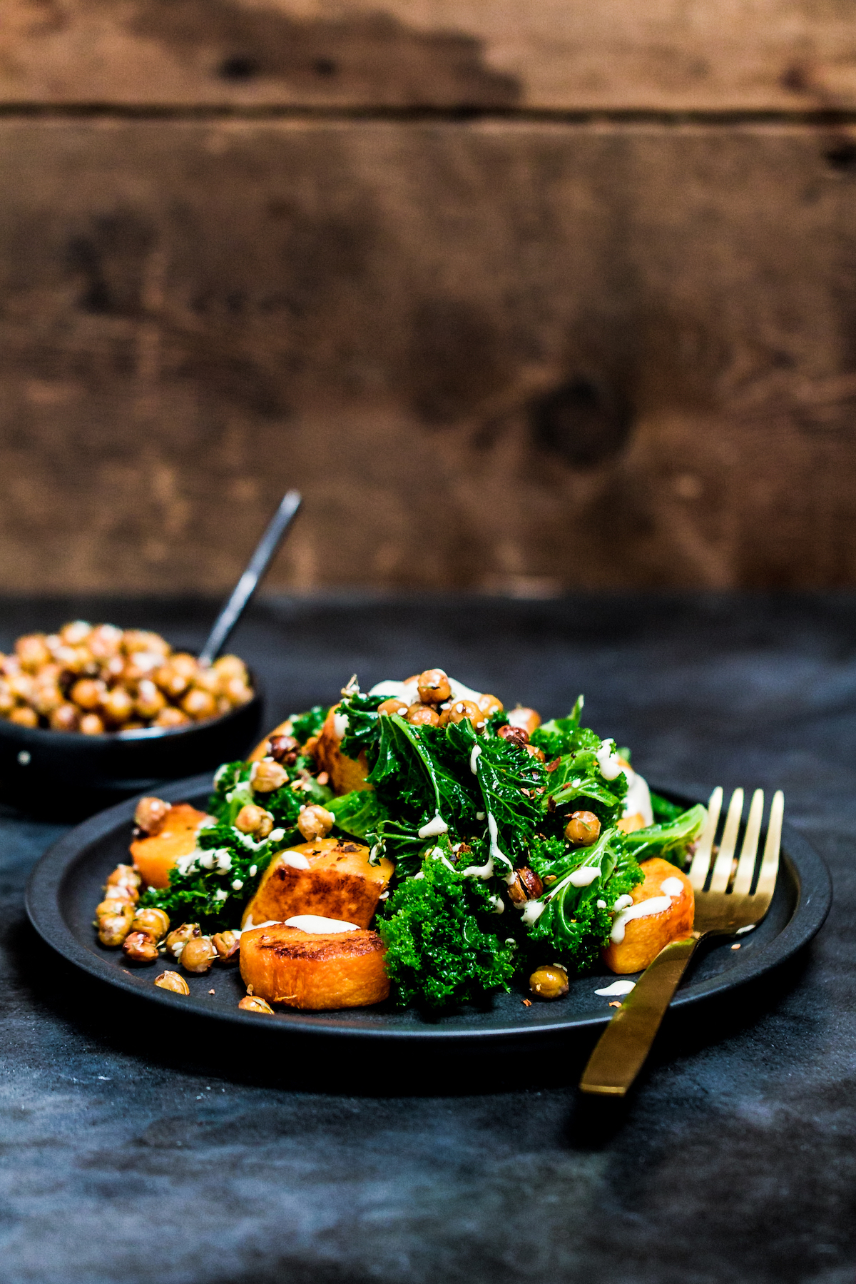Baked butternut squash cubes with steamed kale, crispy chickpeas and tahini sauce on a black plate