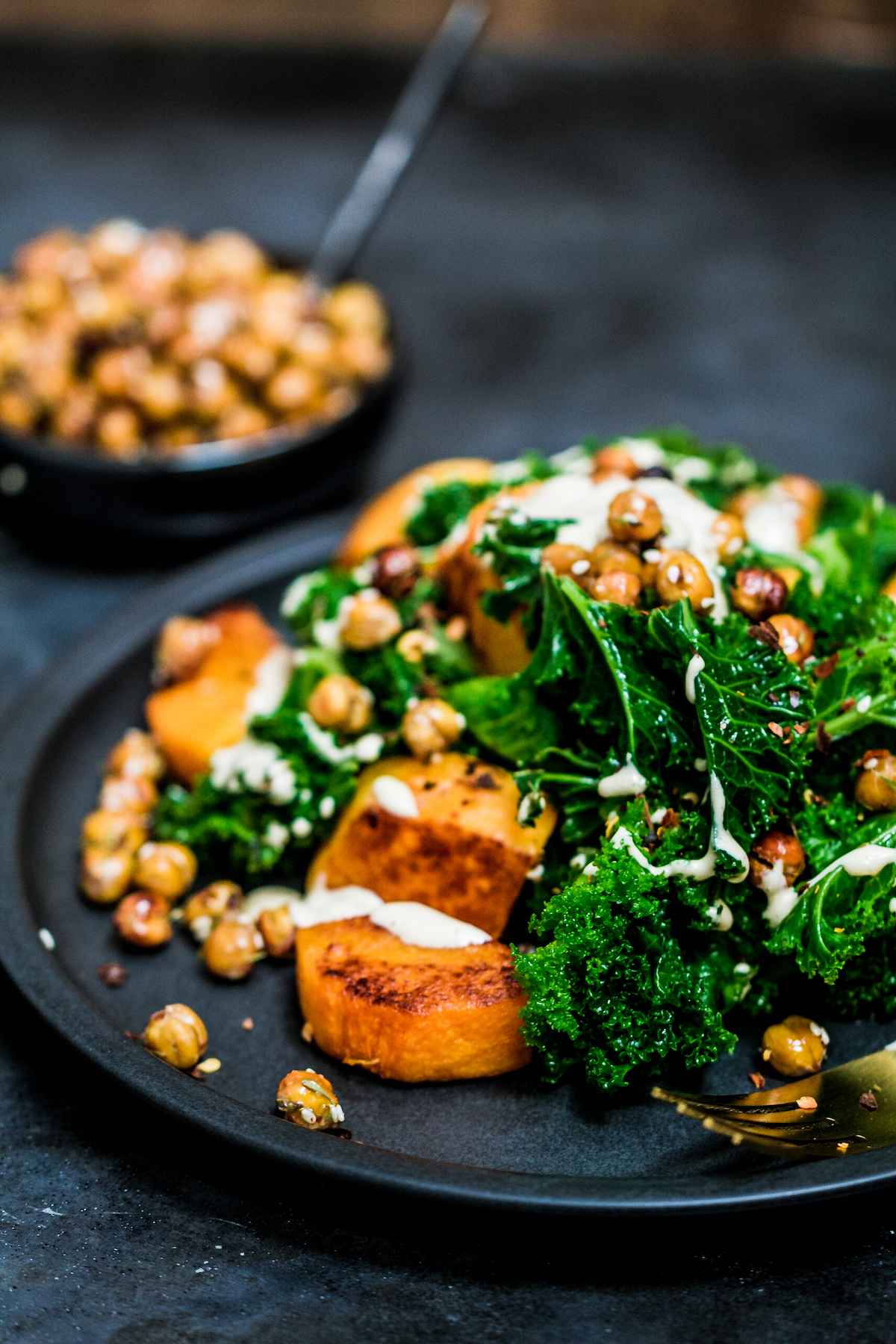 Close up of baked butternut squash cubes with steamed kale, crispy chickpeas and tahini sauce on a black plate