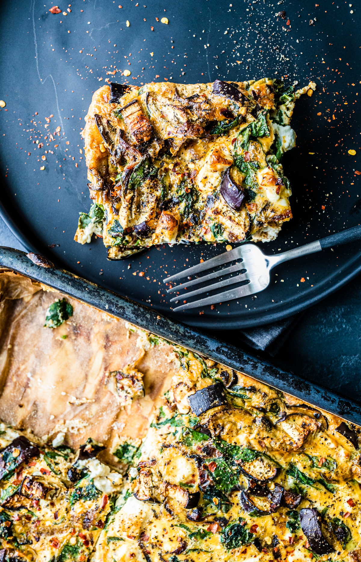 One piece of Caramelised Onion & Feta Frittata from a tray, on a black plate with a fork, top down view.