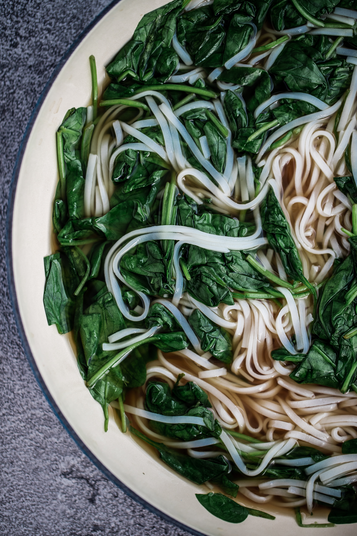 Overhead shot of spinach and white rice noodles in a pan with pho broth
