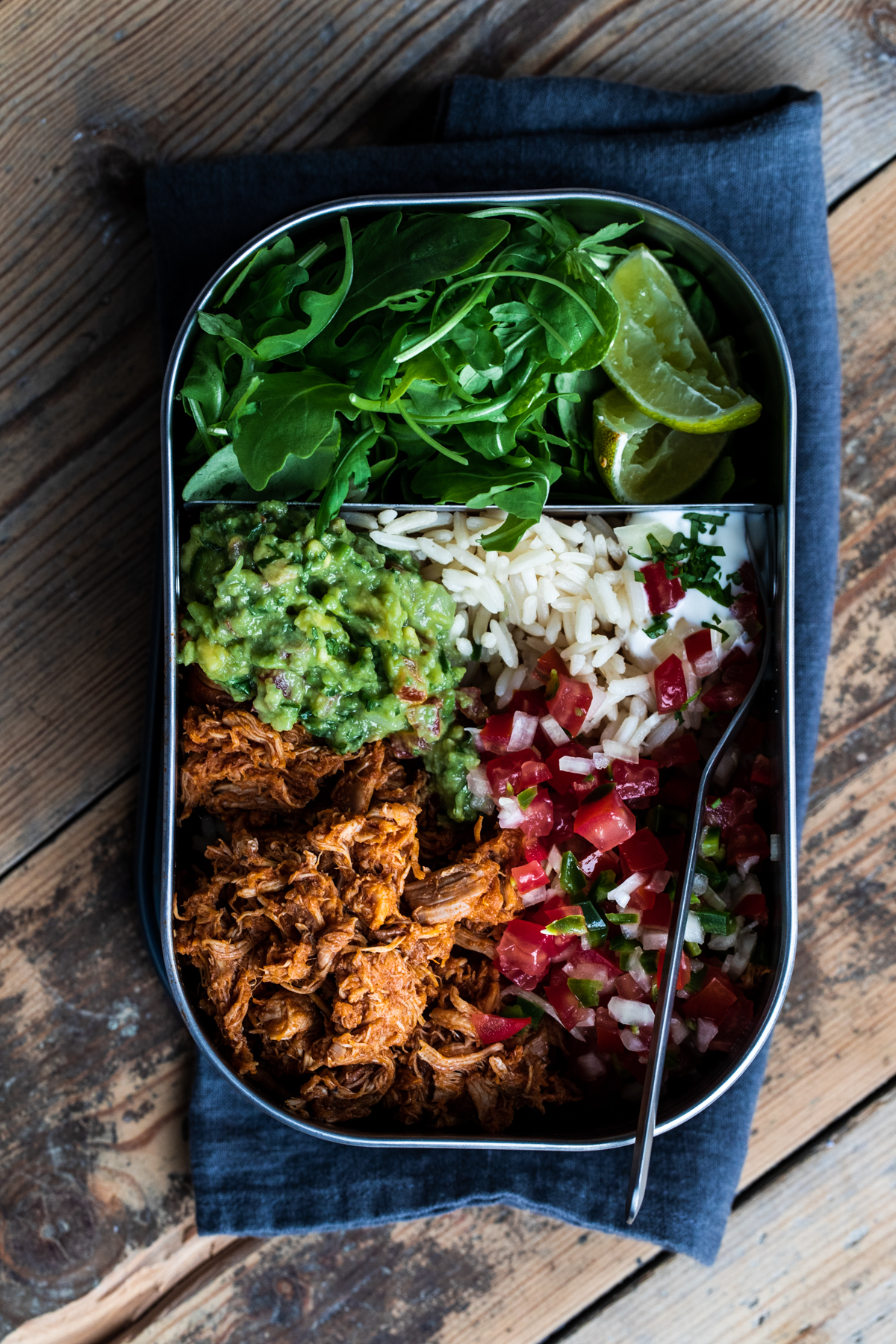 Overhead view of slow cooked chipotle chicken lunch box with guacamole, rice, pico de gallo and greens on a wooden background.