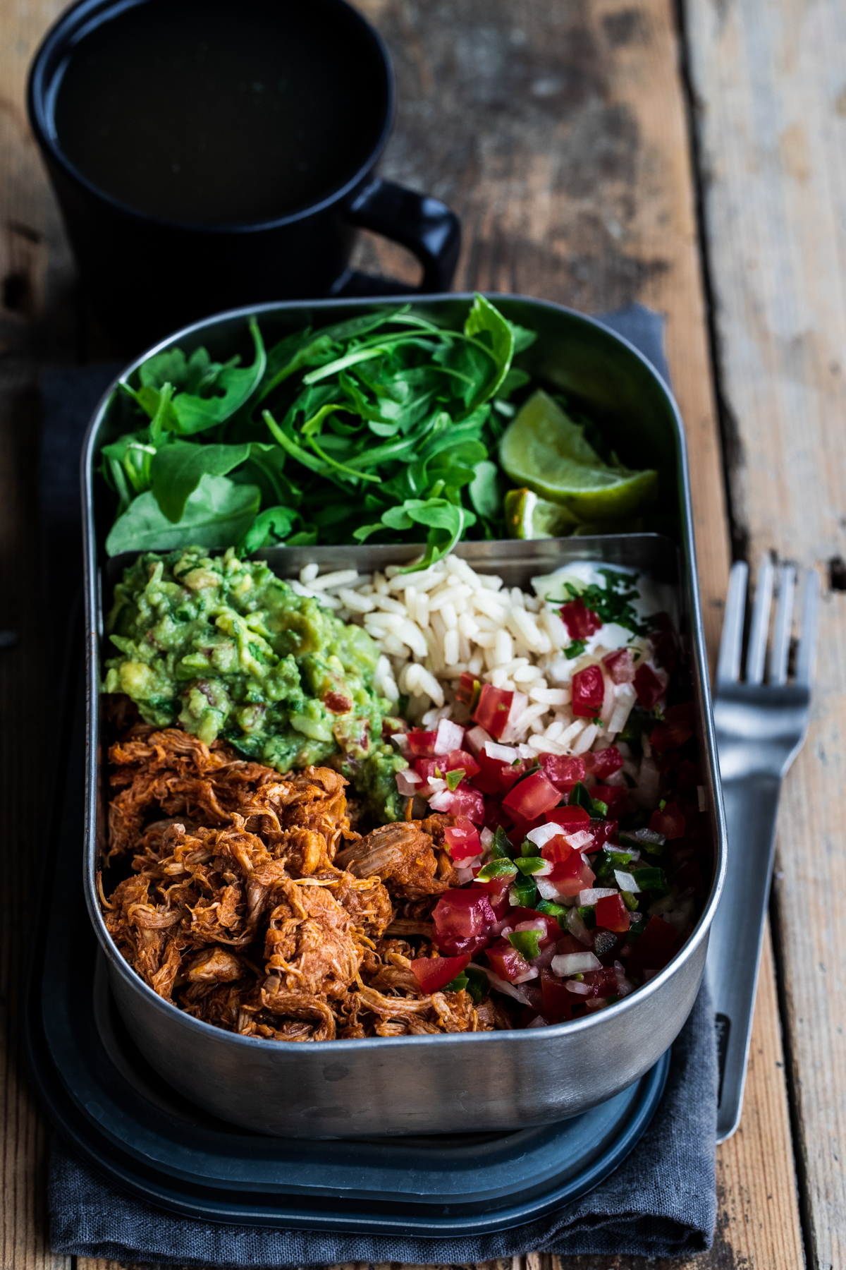 Slow cooked chipotle chicken lunch box with guacamole, rice, pico de gallo and greens on a wooden background.