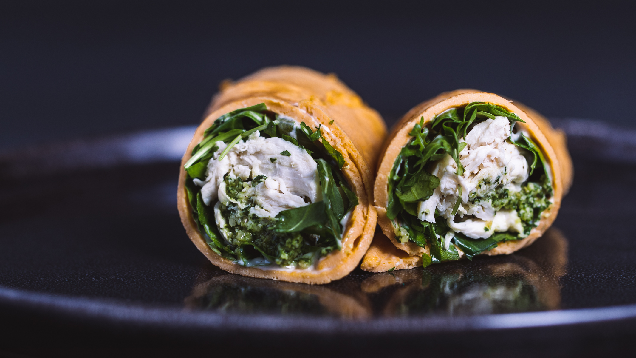 Side view of cut egg wraps with chicken, pesto and arugula on a black plate and background.
