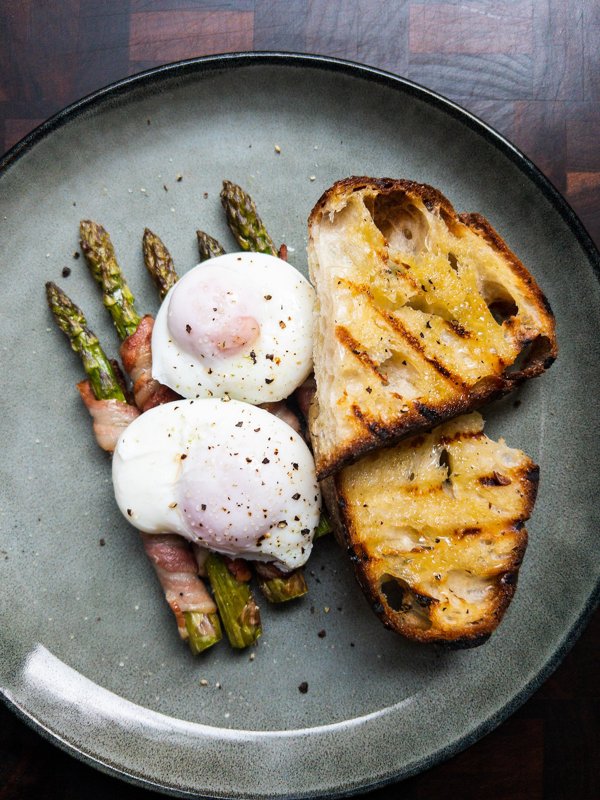 Overhead shot of bacon wrapped asparagus with poached eggs and grilled sourdough on a grey blue plate