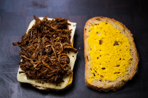 Two pieces of sourdough with mustard, Swiss cheese and pulled pork on a wooden chopping board