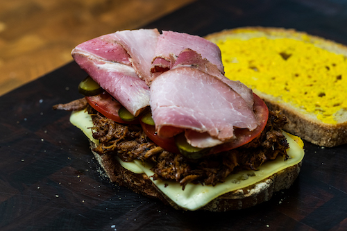 Two pieces of sourdough with mustard, Swiss cheese, pulled pork, tomato, pickles and ham on a wooden chopping board