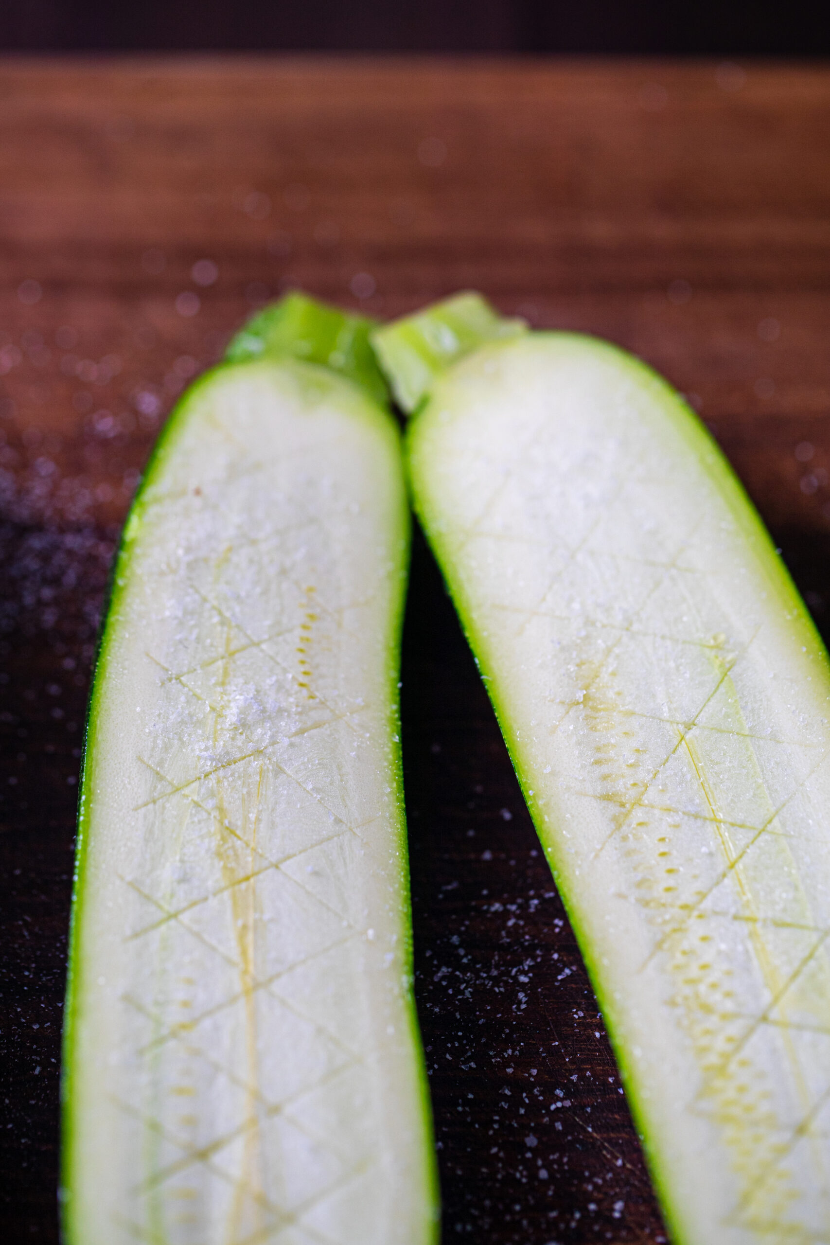 Zucchini scored in a crosshatch pattern and salted, on a wooden chopping board.